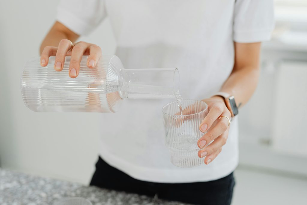 A person pouring water from a carafe into a glass on a countertop indoors.